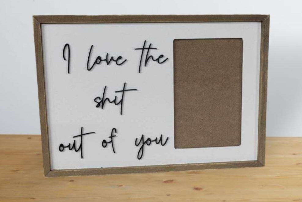 A framed typographic wall sign with the phrase 'I love the shit out of you' in black script on a white background, next to a blank brown rectangular photo slot, leaning on a light wood surface against a white wall with a gray desk lamp partially visible.
